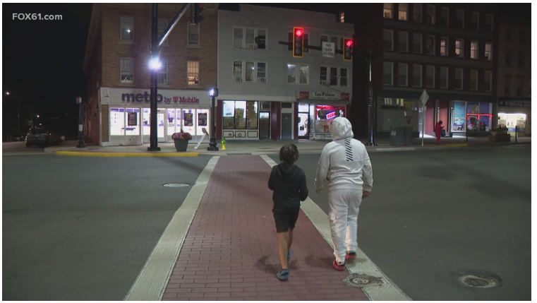 Pedestrians crossing at crosswalk, illuminated by lighting mounted on a traffic signal pole arm.